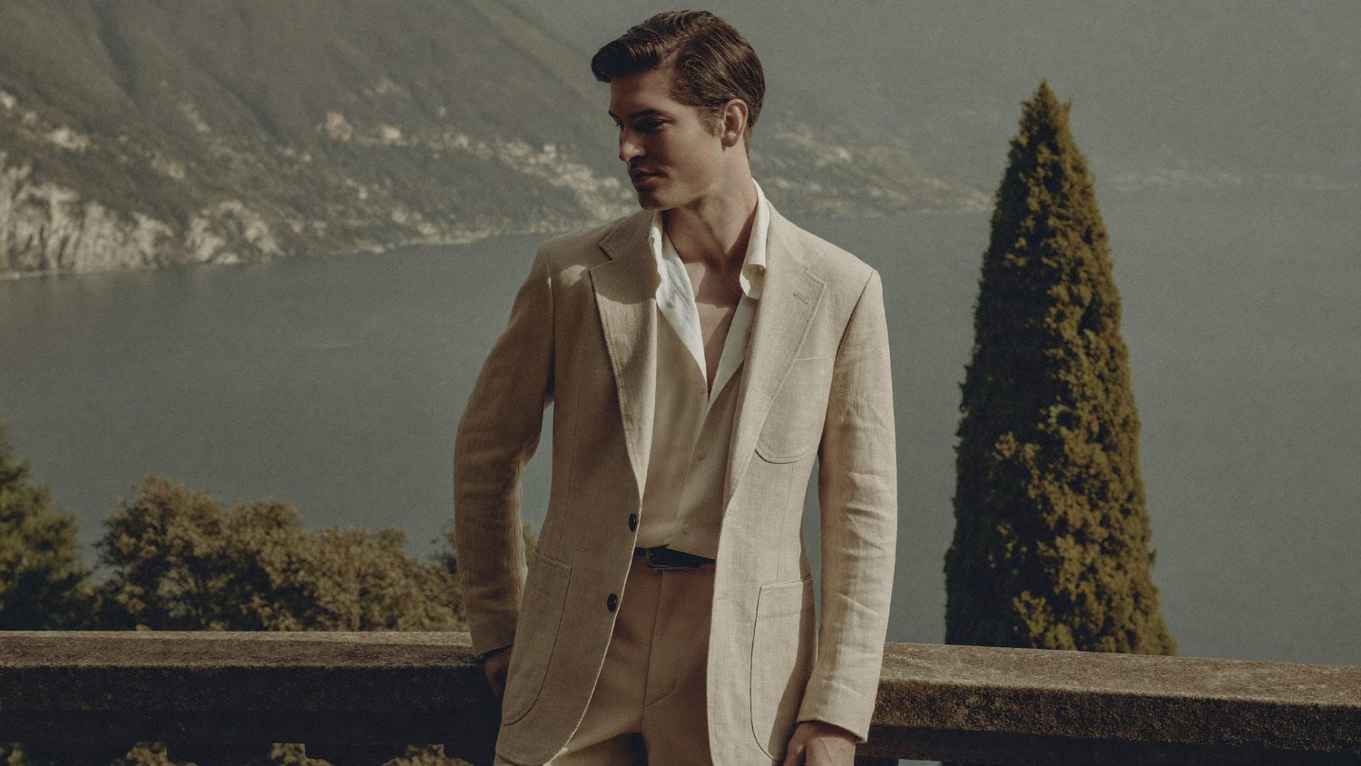 Man in a soft linen jacket on a stone terrace overlooking the lake in morning light