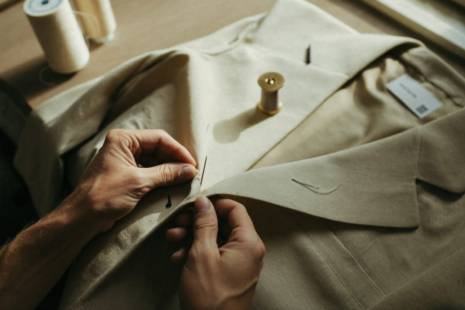 Hands finishing a linen jacket by hand in a sunlit workroom